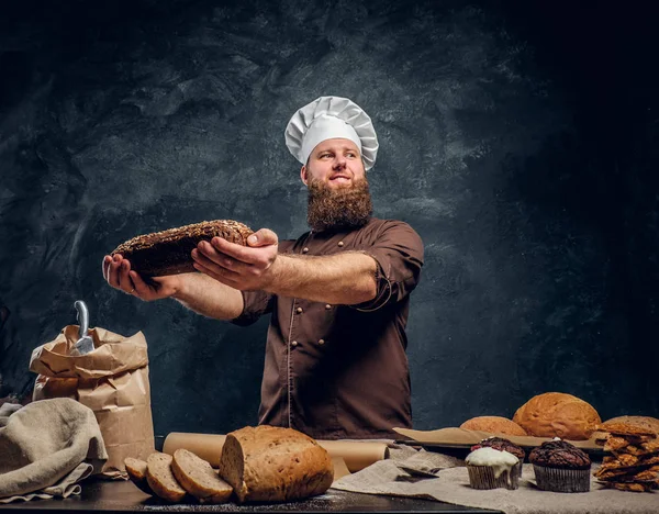 Um padeiro barbudo vestindo um uniforme mostrando pão fresco em pé ao lado de uma mesa, decorado com deliciosos pães, baguetes e muffins — Fotografia de Stock