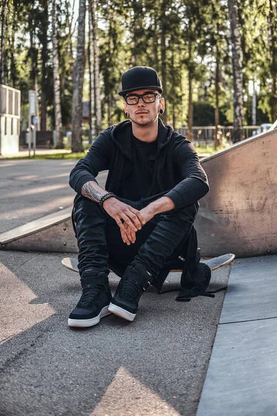 Elegante skateboarder en una sudadera con capucha negra y gorra se sienta en el tablero en el skatepark en la hora de verano — Foto de Stock