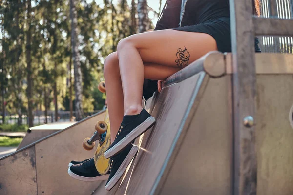 Chicas piernas con un tatuaje. Joven chica elegante sentado en una rampa en el parque de skate en el verano . —  Fotos de Stock