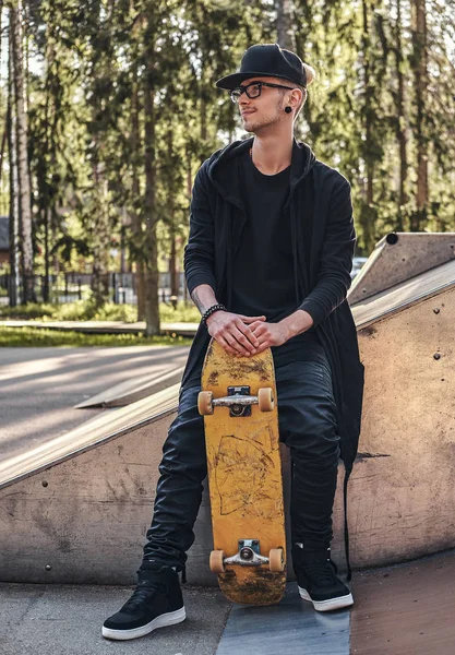 Elegante skateboarder en una sudadera con capucha negra y gorra se sienta en la rampa en el skatepark en la hora de verano — Foto de Stock