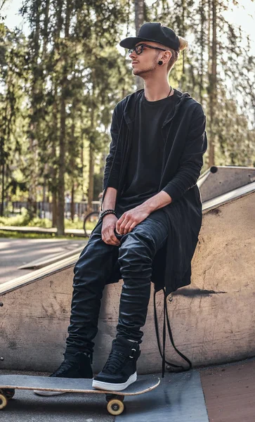 Κομψό skateboarder σε ένα μαύρο hoodie και καπάκι κάθεται στη ράμπα στο skatepark κατά τη θερινή ώρα — Φωτογραφία Αρχείου