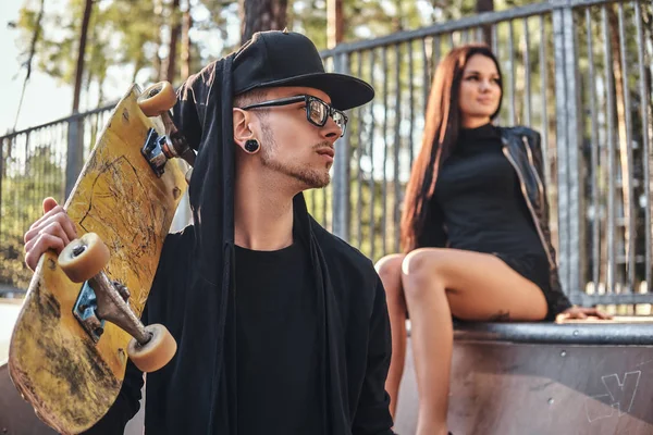 Casal jovem no parque de skate no verão. Retrato de um skate com capuz e chapéu posando com um skate no ombro — Fotografia de Stock