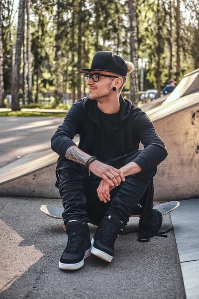 Elegante skateboarder en una sudadera con capucha negra y gorra se sienta en el tablero en el skatepark en la hora de verano —  Fotos de Stock