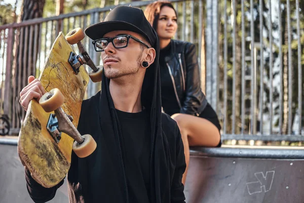 Ungt par i skatepark på sommaren. Porträtt av en skateboard i en hoodie och hatt som poserar med en skateboard på axeln — Stockfoto