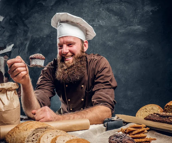 Brodaty baker zadowolony z jego pracy, patrząc na jego świeżo wykonane muffin jednocześnie opierając się na stole w ciemnym studio — Zdjęcie stockowe