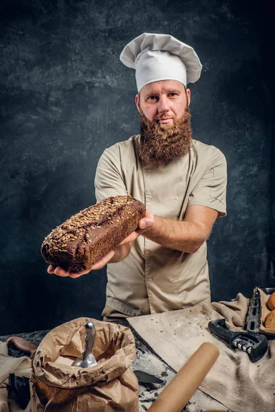 Um padeiro barbudo vestindo um uniforme mostrando pão fresco em pé ao lado de uma mesa, decorado com deliciosos pães, baguetes — Fotografia de Stock