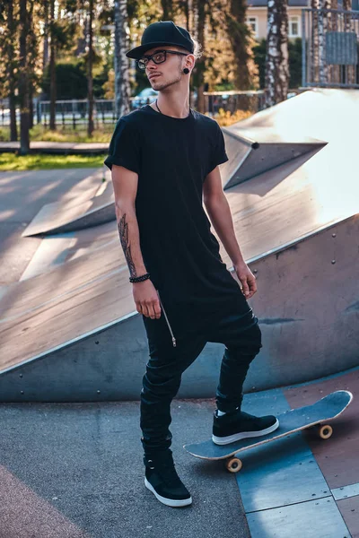 Κομψό skateboarder σε ένα μαύρο πουκάμισο και το καπάκι που στέκεται πάνω σε ένα Διοικητικό Συμβούλιο στο skatepark κατά την καλοκαιρινή περίοδο — Φωτογραφία Αρχείου