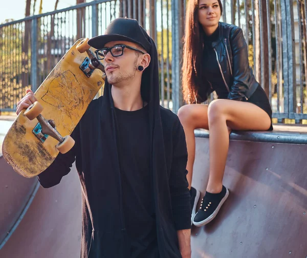 Ungt par i skatepark på sommaren. Porträtt av en skateboard i en hoodie och hatt som poserar med en skateboard på axeln — Stockfoto