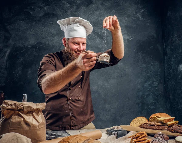 Alegre padeiro barbudo polvilhando um pouco de farinha em seu queque acabado de fazer ao lado de uma mesa — Fotografia de Stock
