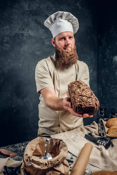 Um padeiro barbudo vestindo um uniforme mostrando pão fresco em pé ao lado de uma mesa, decorado com deliciosos pães, baguetes — Fotografia de Stock