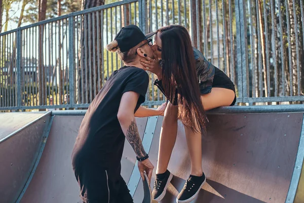 Стильная влюбленная пара. Красивая девушка сидит на рампе и целует своего парня в скейтпарке летом — стоковое фото