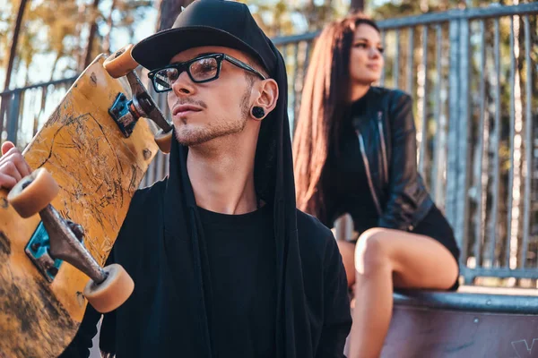 Jeune couple dans le skatepark en été. Portrait d'une planche à roulettes en sweat à capuche et chapeau posant avec une planche à roulettes sur l'épaule — Photo