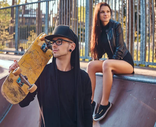 Jeune couple dans le skatepark en été. Portrait d'une planche à roulettes en sweat à capuche et chapeau posant avec une planche à roulettes sur l'épaule — Photo