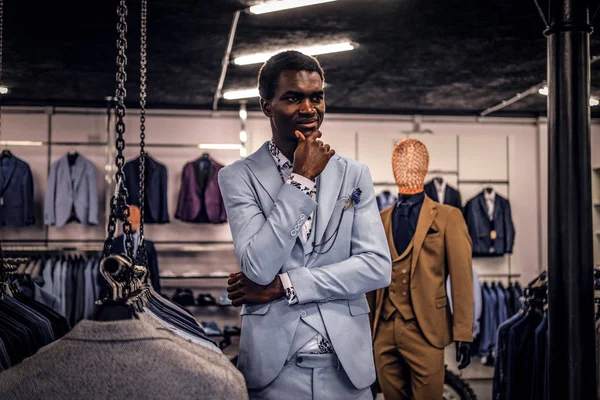 Красивый элегантно одетый афроамериканец позирует с рукой на подбородке с вдумчивым взглядом, стоя в классическом магазине мужской одежды . — стоковое фото