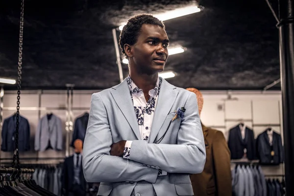 African-American man dressed in blue elegant suit posing with crossed arms in a classic menswear store.