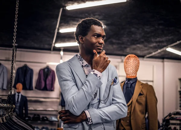 Un hombre afroamericano guapo elegantemente vestido posando con la mano en la barbilla con una mirada reflexiva mientras está de pie en una tienda de ropa masculina clásica . — Foto de Stock