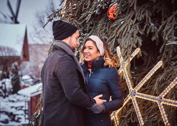Casal feliz vestindo roupas quentes de mãos dadas e olhar um para o outro, de pé perto de uma árvore de Natal da cidade, gostando de passar o tempo juntos . — Fotografia de Stock