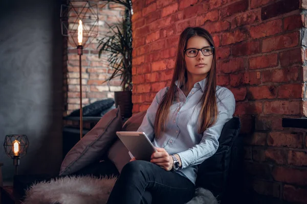 Hermosa mujer joven vestida con camisa elegante sostiene una tableta, se sienta en el sofá en una habitación con un interior loft y mirando hacia otro lado — Foto de Stock