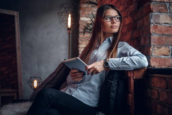 Hermosa mujer joven vestida con camisa elegante sostiene una tableta, se sienta en el sofá en una habitación con un interior loft y mirando hacia otro lado — Foto de Stock
