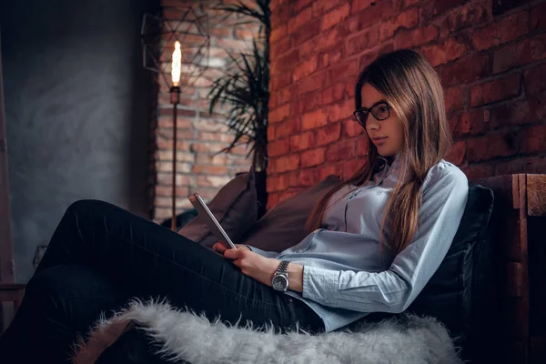 Hermosa joven vestida con camisa elegante que trabaja en una tableta, se sienta en el sofá en una habitación con un interior loft — Foto de Stock