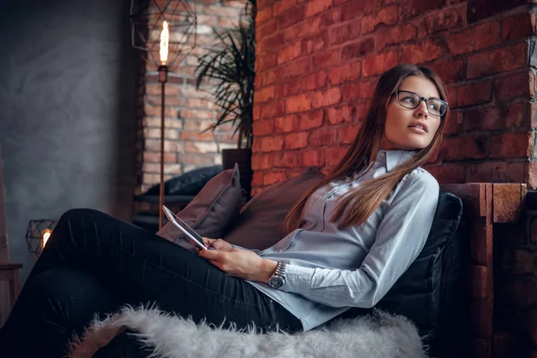 Красивая молодая женщина в элегантной рубашке держит планшет, сидит на диване в комнате с интерьером лофта и смотрит в сторону — стоковое фото