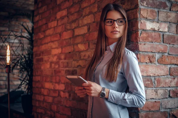 Умная девушка, одетая в элегантную рубашку, держащая планшет, опираясь на кирпичную стену в комнате с интерьером лофта — стоковое фото