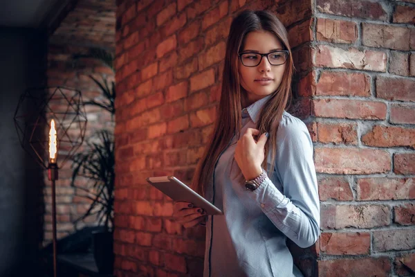 Chica inteligente vestida con una camisa elegante sosteniendo la tableta mientras se apoya en una pared de ladrillo en la habitación con un interior loft — Foto de Stock