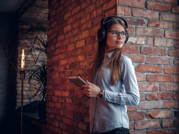 Chica en camisa elegante sosteniendo una tableta disfruta de música favorita en los auriculares inalámbricos de pie cerca de la pared de ladrillo — Foto de Stock