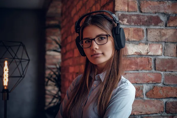 Chica con encanto en camisa elegante disfruta de música favorita en los auriculares inalámbricos de pie cerca de la pared de ladrillo — Foto de Stock