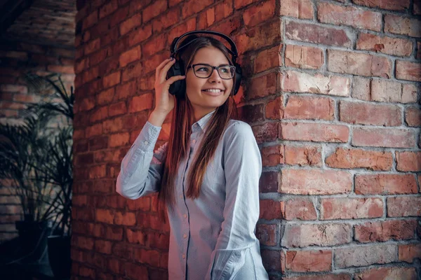 Menina encantadora na camisa elegante goza de música favorita em fones de ouvido sem fio de pé perto da parede de tijolo — Fotografia de Stock