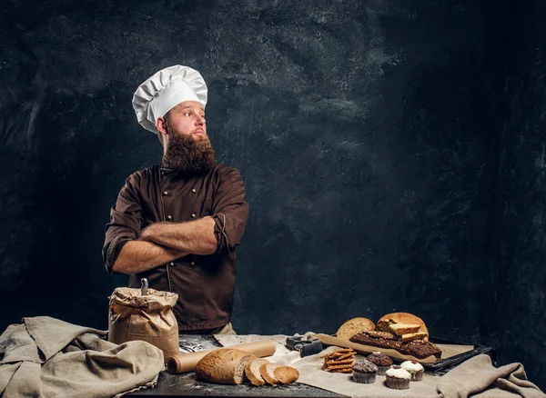 Um padeiro barbudo vestindo um uniforme de pé com os braços cruzados ao lado de uma mesa, decorado com deliciosos pães, baguetes e muffins — Fotografia de Stock