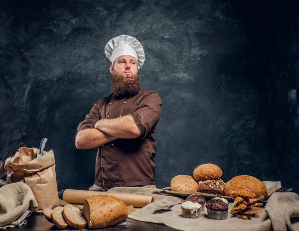 Um padeiro barbudo vestindo um uniforme de pé com os braços cruzados ao lado de uma mesa, decorado com deliciosos pães, baguetes e muffins — Fotografia de Stock