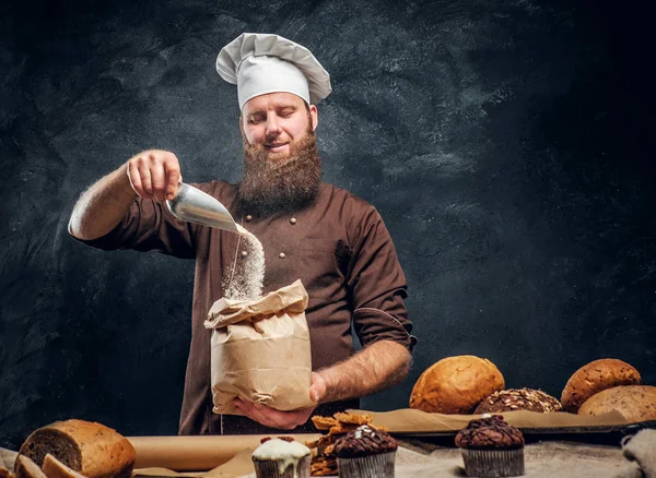 Brodaty baker, noszenie munduru gotować niektóre pierwotności w torbie, stojący obok tabeli, ozdobione pyszne chleby, bagietki — Zdjęcie stockowe