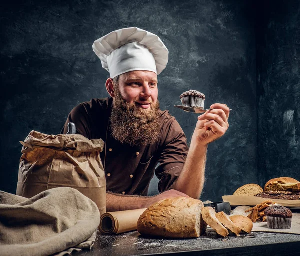Brodaty baker zadowolony z jego pracy, patrząc na jego świeżo wykonane muffin jednocześnie opierając się na stole w ciemnym studio — Zdjęcie stockowe