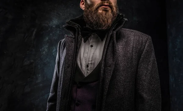 Levágott kép egy elegáns szakállas férfi öltözve egy elegáns ruha, és a kabát állt ellen a sötét mintás fal — Stock Fotó