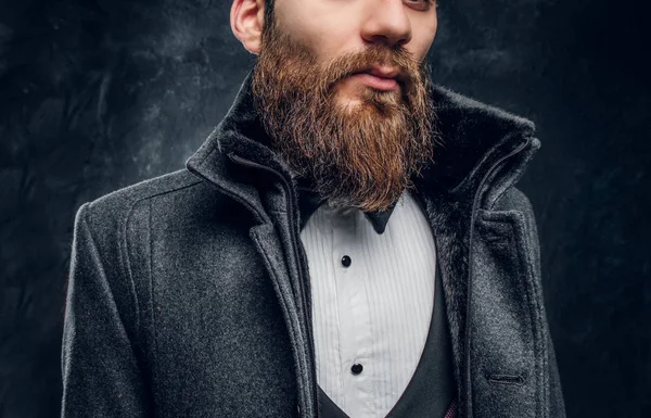 Cropped image of a stylish bearded man dressed in an elegant suit and coat standing against a dark textured wall — Stock Photo, Image