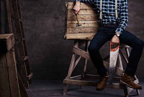 Cropped image. Worker, carpenter, handyman man wearing a checkered shirt with suspenders holding a hammer and saw while sitting on a wooden scaffolding