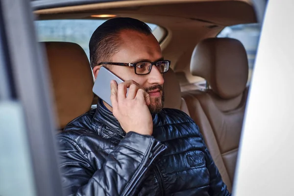 Homem de negócios na moda vestido sentado em um banco de trás no carro de luxo, falando por um telefone — Fotografia de Stock