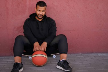 Afrikalı-Amerikalı adam bir kaykay üzerinde oturan ve bir basketbol dışında bir duvara yaslanmış Ise tutarak siyah kapüşonlu