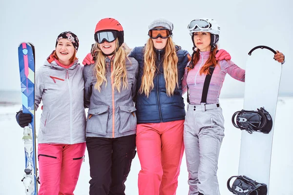 Mulheres amigas em esportes roupas de inverno com snowboards e esquis de pé juntos em um abraço e olhando para a câmera em uma praia nevada — Fotografia de Stock