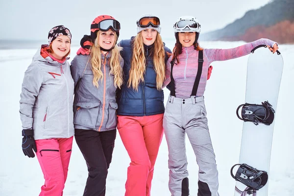 Women friends in sports winter clothes with snowboards and skis standing together in a hug and looking at the camera at a snowy beach