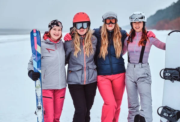 Women friends in sports winter clothes with snowboards and skis standing together in a hug and looking at the camera at a snowy beach