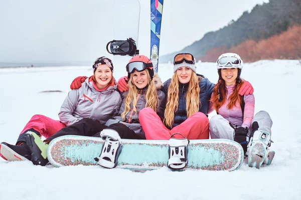 Γυναίκες φίλους σε σπορ χειμερινά ρούχα με σνόουμπορντ και σκι αγκάλιασμα μαζί και να βλέπουν μια φωτογραφική μηχανή, ενώ κάθεται σε μια χιονισμένη παραλία — Φωτογραφία Αρχείου