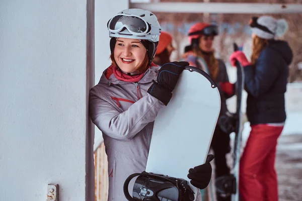 Mujer sonriente con ropa deportiva de invierno posando con una tabla de snowboard en la estación de esquí de invierno —  Fotos de Stock