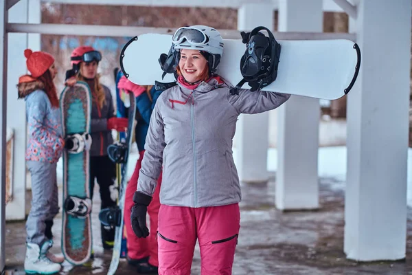 Mujer sonriente con ropa deportiva de invierno posando con una tabla de snowboard en la estación de esquí de invierno —  Fotos de Stock
