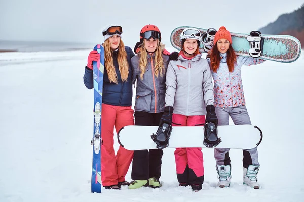 Women friends in sports winter clothes with snowboards and skis standing together in a hug and looking at the camera