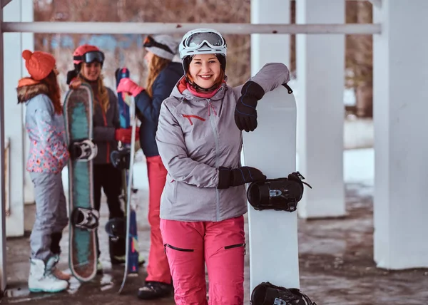 Mujer sonriente con ropa deportiva de invierno posando con una tabla de snowboard en la estación de esquí de invierno —  Fotos de Stock