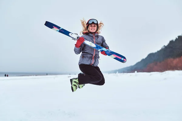 Mujer alegre usando ropa deportiva de calentamiento saltando con esquís en una playa nevada —  Fotos de Stock