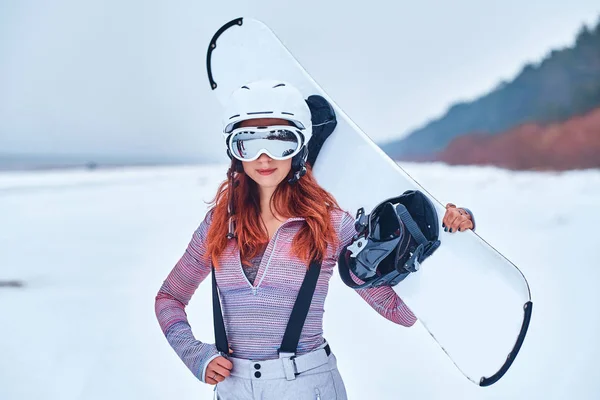 Porträtt av en vacker rödhårig flicka i skyddande hjälm och glasögon håller en snowboard, poserar på en snöig strand — Stockfoto