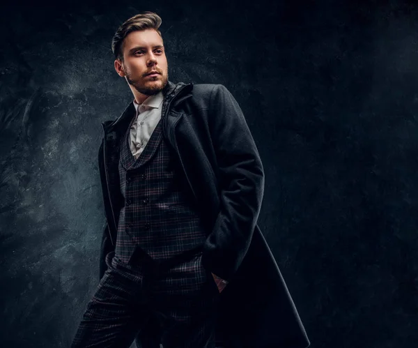 A stylish man dressed in an elegant suit and coat posing with a hand in a pocket against a dark textured wall